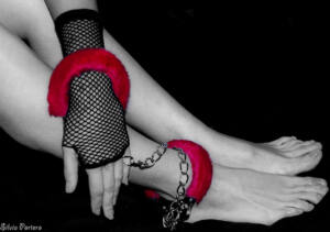 Foto en blanco y negro de una mano enfundada en guante de rejilla esposada a su propio tobillo por una esposa de terciopelo roja