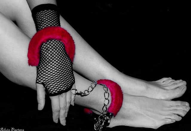 Foto en blanco y negro de una mano enfundada en guante de rejilla esposada a su propio tobillo por una esposa de terciopelo roja