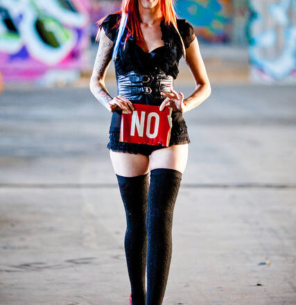 Chica pelirroja con minifalda, medias negras hasta el muslo con un cartel en el que se lee: NO