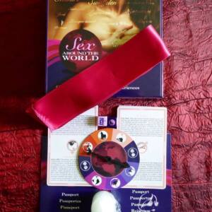 Foto con el contenido de sexo en todo el mundo: cinta de seda rosa sobre la caja del juego, ruleta sobre las castas/visados. Encima de la ruleta los dos dados: uno rosa y otro morado. Debajo de la ruleta, una velita blanca