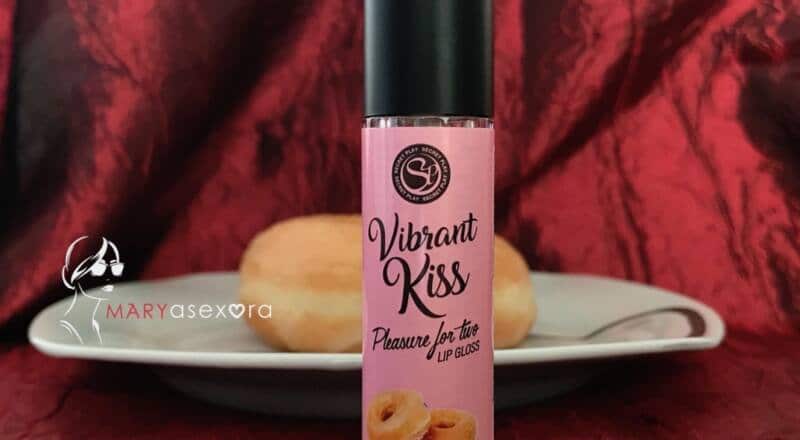 Aparece el Lip gloss vibrant kiss sabor donut en primer plano y detrás un plato con un donut glaseado.