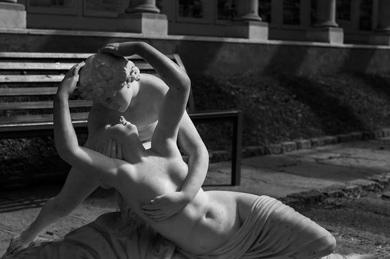 Estatua de mármol con una mujer recostada con el torso desnudo y un hombre la abraza y besa desde atrás