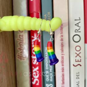 Pendientes dildos arcoíris. Pendientes con forma de dildo hechos con fimo formando los colores del arcoíris colgando sobre un dildo de juguete en la estantería de libros.