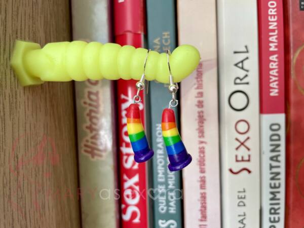 Pendientes dildos arcoíris. Pendientes con forma de dildo hechos con fimo formando los colores del arcoíris colgando sobre un dildo de juguete en la estantería de libros.