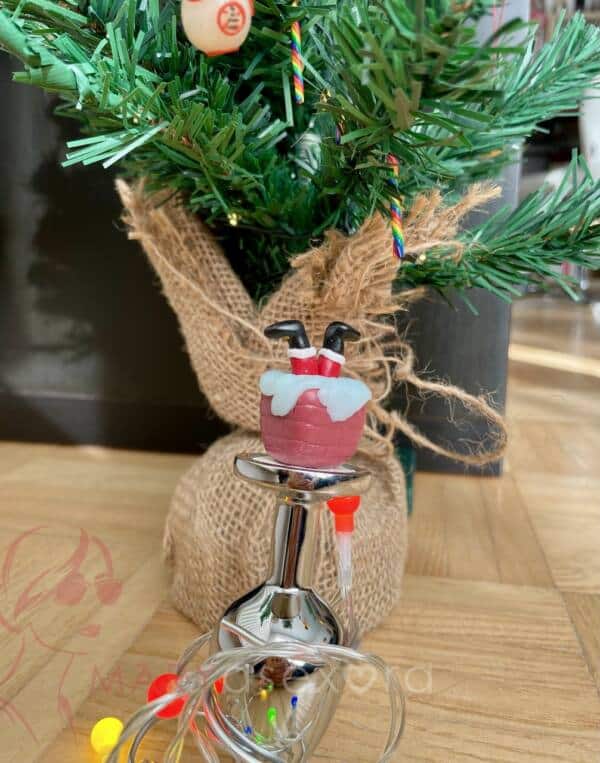 Plug navideño: plug de acero en cuya base hay una chimenea y unas piernas de Papá Noél hechos con Fimo. Detrás del plug, un árbol de Navidad.