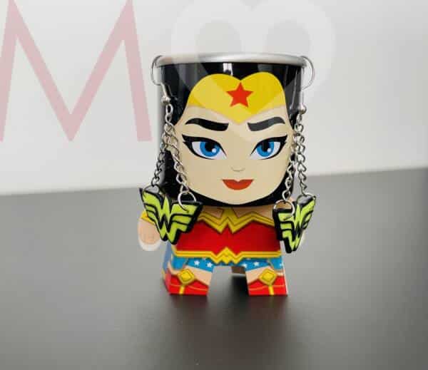 Pendientes logotipo Wonder Woman hechos a mano con polimer clay amarillo efecto neón y negro puestos en una figura de Wonder Woman con cuerpo de papel y cabeza de lata.