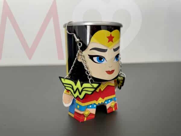 Pendientes logotipo Wonder Woman hechos a mano con polimer clay amarillo efecto neón y negro puestos en una figura de Wonder Woman con cuerpo de papel y cabeza de lata.